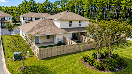 A house with a brown roof and a white exterior is surrounded by a wooden fence and green bushes.