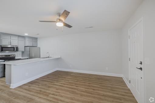 A room with a ceiling fan and a kitchen area with white cabinets.