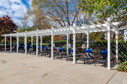 A white pergola with blue chairs is in the foreground of a sunny day.
