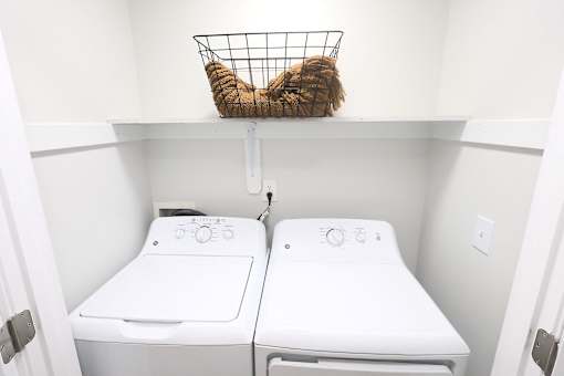 A laundry basket with a cat in it sits on top of a shelf above two washing machines.