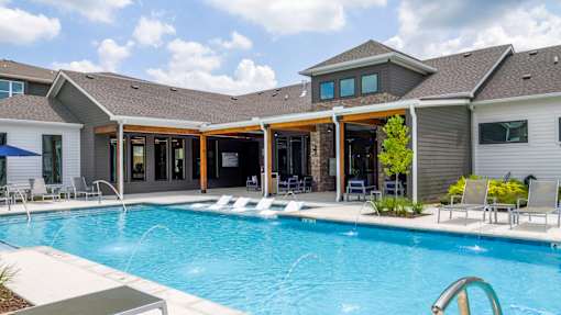A large swimming pool in front of a house with a patio and lounge chairs.