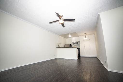 an empty living room with a ceiling fan and a kitchen in the background