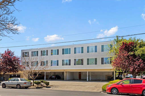 A red car is parked in front of a white building with a parking lot in front.