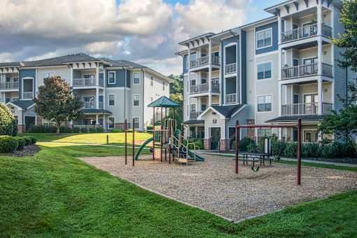 A playground area with a slide, swings, and a seesaw in front of apartment buildings.