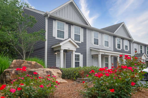 A house with grey siding and white trim is surrounded by red flowers and greenery.