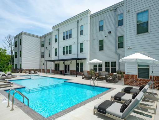 One Bedroom Apartments in Lakeland TN - Lakeland Town Square - Pool Area with Multiple Levels, Lounge Chairs with Cushions, white Umbrellas, Seating Area with Table, Brick Details, and Nearby Foliage