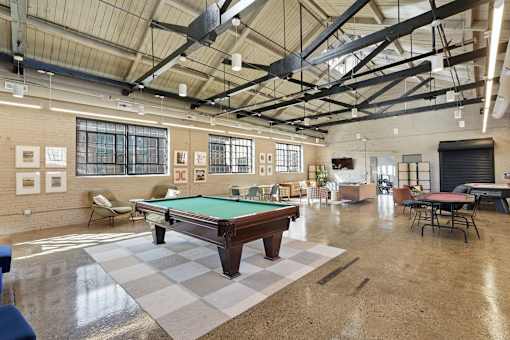 A pool table is in the middle of a room with a high ceiling and lots of windows.