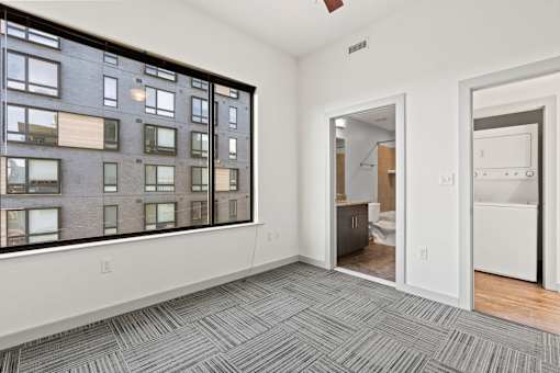 A room with a grey carpet and a large window overlooking a building.