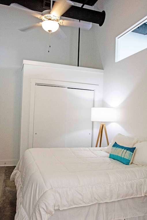 A bed with a white comforter and a blue pillow is in a room with a ceiling fan.
