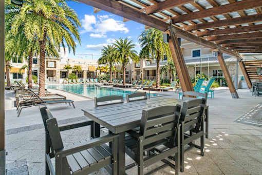 A wooden table and chairs are set up under a pergola by a pool. at Centre Pointe Apartments, Melbourne, FL, 32940