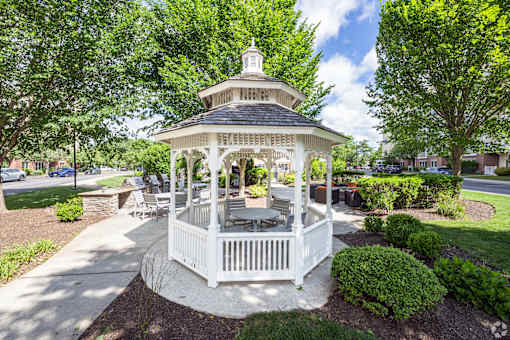 A gazebo with a white railing and a black roof is surrounded by green bushes and trees.