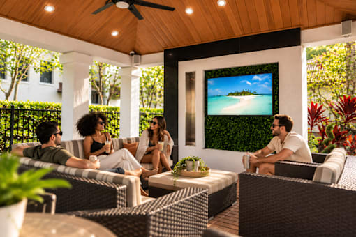 A group of people are sitting on a patio with a TV screen showing a beach scene.
