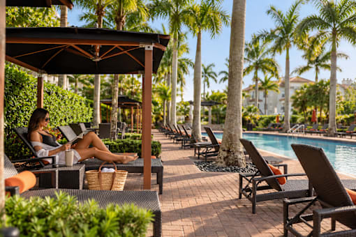 A woman is sitting on a lounge chair by a pool, reading a book.