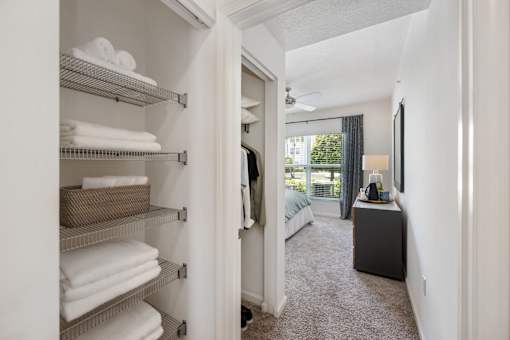 A white closet with towels on shelves and a bedroom with a bed and a dresser in the background.