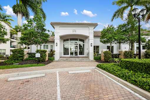 The image shows a white building with a glass entrance and a brick pathway leading to it.