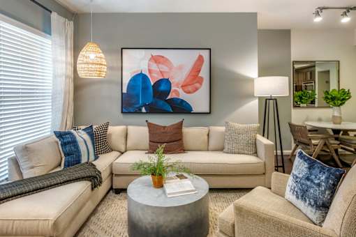 A living room with a large abstract painting on the wall, a beige sofa, a grey coffee table, and a dining table with chairs.