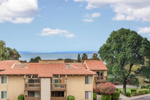 A building with a red tile roof is surrounded by greenery and a body of water.