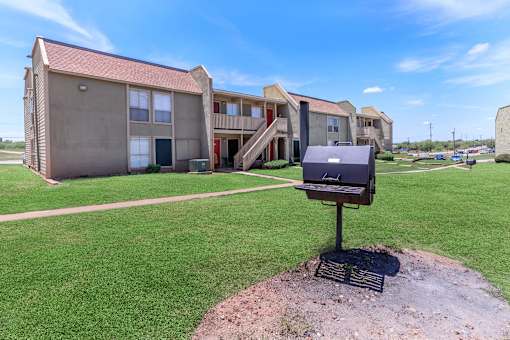A barbecue grill is in the foreground of a grassy area in front of apartment buildings.