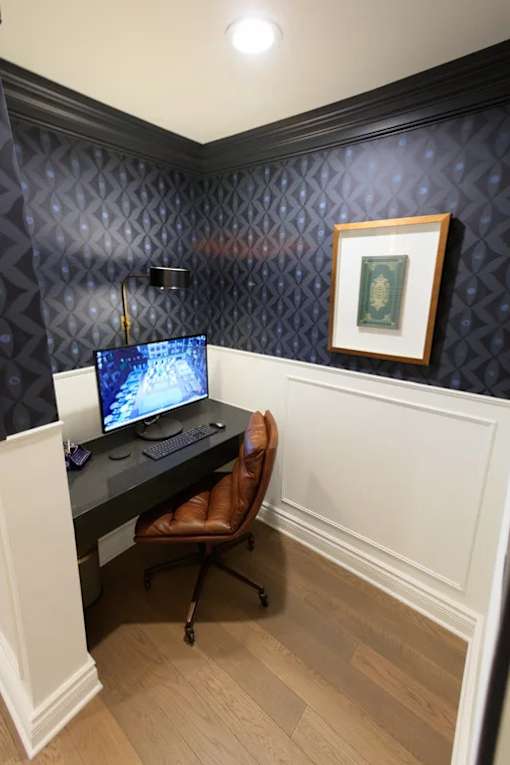 A computer desk with a monitor, keyboard, and mouse is set up in a corner with a chair and a picture frame on the wall.