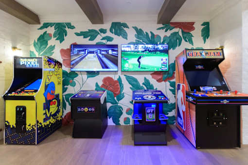 A row of arcade game machines are lined up against a wall with a leafy mural.