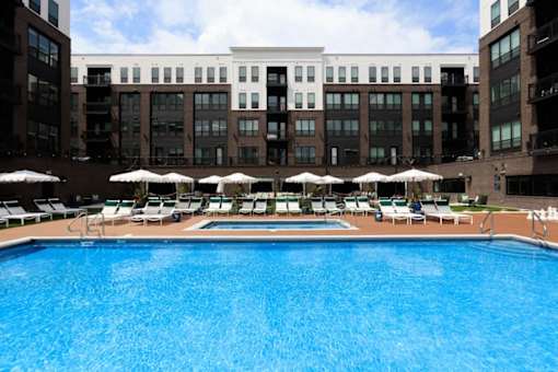 A large swimming pool surrounded by lounge chairs and umbrellas in front of a multi-story building.