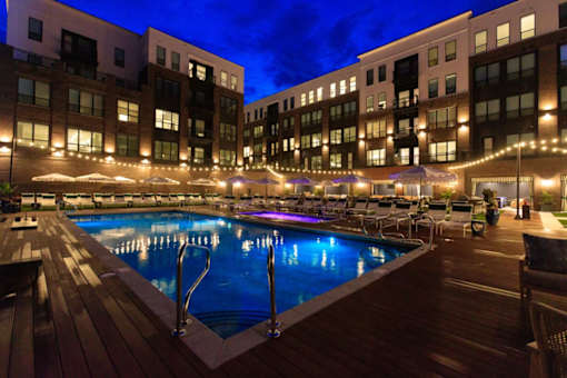 A pool area with a wooden deck and chairs is lit up at night.