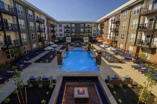A large swimming pool surrounded by lounge chairs and umbrellas in a courtyard surrounded by apartment buildings.