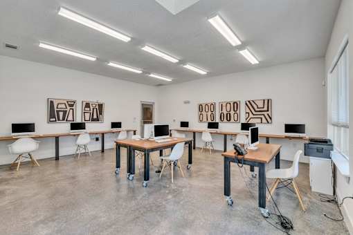 A room with white walls and a concrete floor has several desks with computers on them.