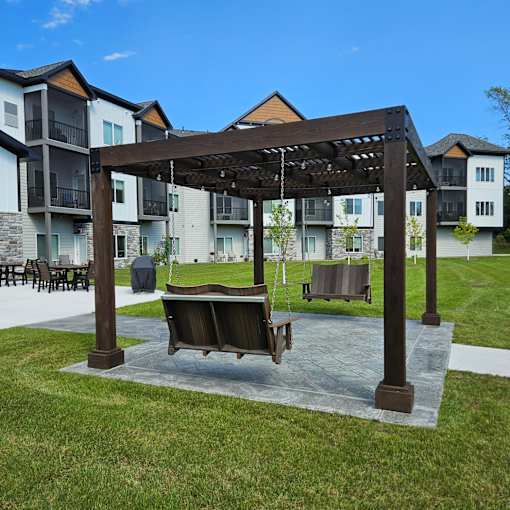 A wooden pergola with a hanging bench is in the foreground of a grassy area with apartment buildings in the background.