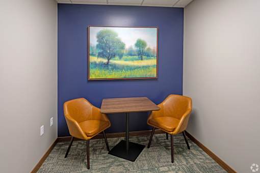 A painting of a tree in a field hangs on a blue wall above a table with two chairs.