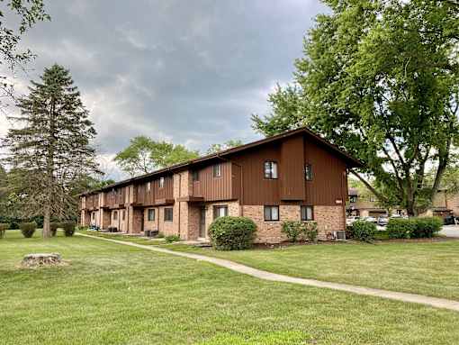 A long building with a brown roof and windows is surrounded by trees and grass at Root River Apartments, Greendale, WI, 53129