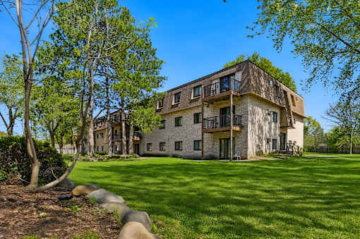 A large, two-story house with a balcony on the second floor is surrounded by a grassy lawn and trees.