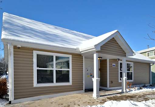 A small house with a brown siding and a white trim is covered in snow.