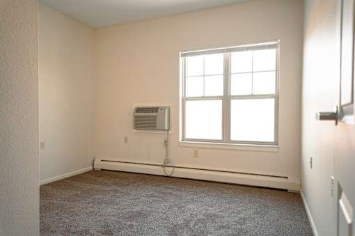 A room with a window, carpet, and a wall-mounted air conditioner unit.