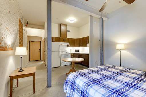 A bedroom with a bed, a table, and a kitchen area in the background.