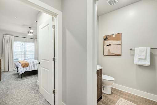 A white bathroom with a toilet, sink, and a bedroom with a bed and window at The Ashton at East Park Apartments, Kennesaw, Georgia