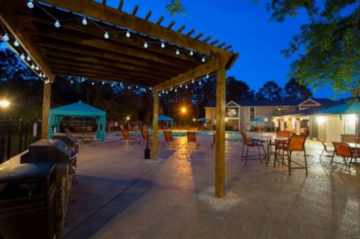 A patio area with a pergola and chairs is lit up at night.