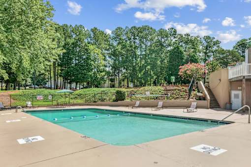 A large outdoor swimming pool surrounded by trees and a patio area with chairs and a table at Emerald Bay, Charlotte, NC, 28217