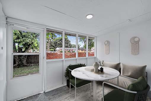 A white table with a vase and a plant on it sits in front of a window with a view of a red fence and trees.