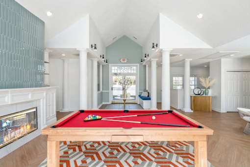 A pool table with a red cloth and white cues is in the middle of a room with a fireplace on the left and a chair on the right.