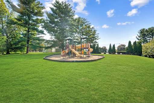 A playground with a round swing set surrounded by a green lawn and trees.
