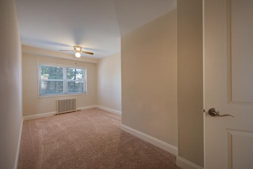 A room with a carpeted floor, a ceiling fan, and a window with blinds.