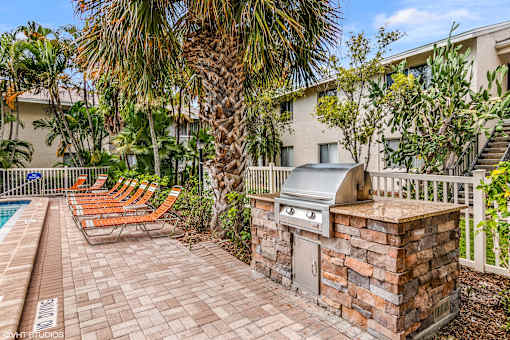 A patio with a grill and chairs is surrounded by a pool and trees. at Sarasota South Apartments, Florida