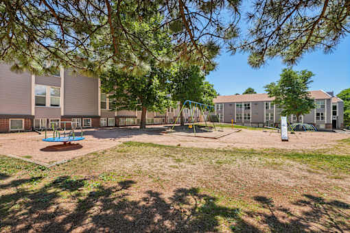 A playground area with a swing set and a slide is surrounded by trees and a building.