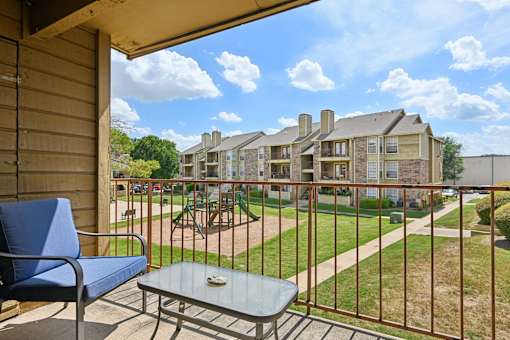 A balcony with a blue chair and table overlooks a grassy area and buildings.