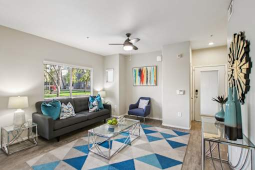 A living room with a grey couch, a blue chair, and a glass coffee table at Sweetwater At Metro North Apartments, Phoenix, AZ