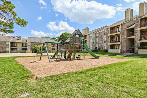 A playground with a green slide and sandbox in front of apartment buildings.