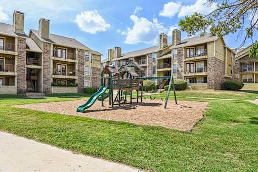 A playground with a green slide and a brown sandbox in front of apartment buildings.