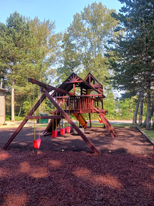 A wooden swing set with red swings is in the foreground of a park.