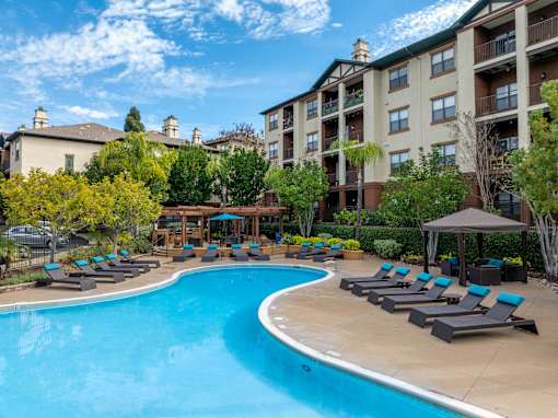 A swimming pool surrounded by lounge chairs and umbrellas in front of apartment buildings.